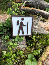 The sign features a simplistic, yet striking, icon of a person with a backpack and walking stick, gracefully representing the journey into hiking. This is rendered in crisp brown on a clean white background, making it easy to pair with any style of decor.
