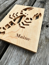 Wood trivet with a lobster cut out of it and the word Maine engraved on the bottom right corner.