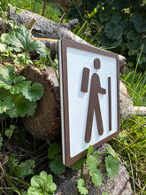 The sign features a simplistic, yet striking, icon of a person with a backpack and walking stick, gracefully representing the journey into hiking. This is rendered in crisp brown on a clean white background, making it easy to pair with any style of decor.
