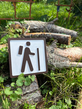 The sign features a simplistic, yet striking, icon of a person with a backpack and walking stick, gracefully representing the journey into hiking. This is rendered in crisp brown on a clean white background, making it easy to pair with any style of decor.
