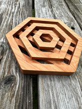 A hexagon shaped wood trivet with hexagon shapes  cut out of it sits on a grey wood table.