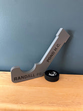 Gray ice hockey stick engraved with team name and player name and number , with puck sitting next to it on a wood table